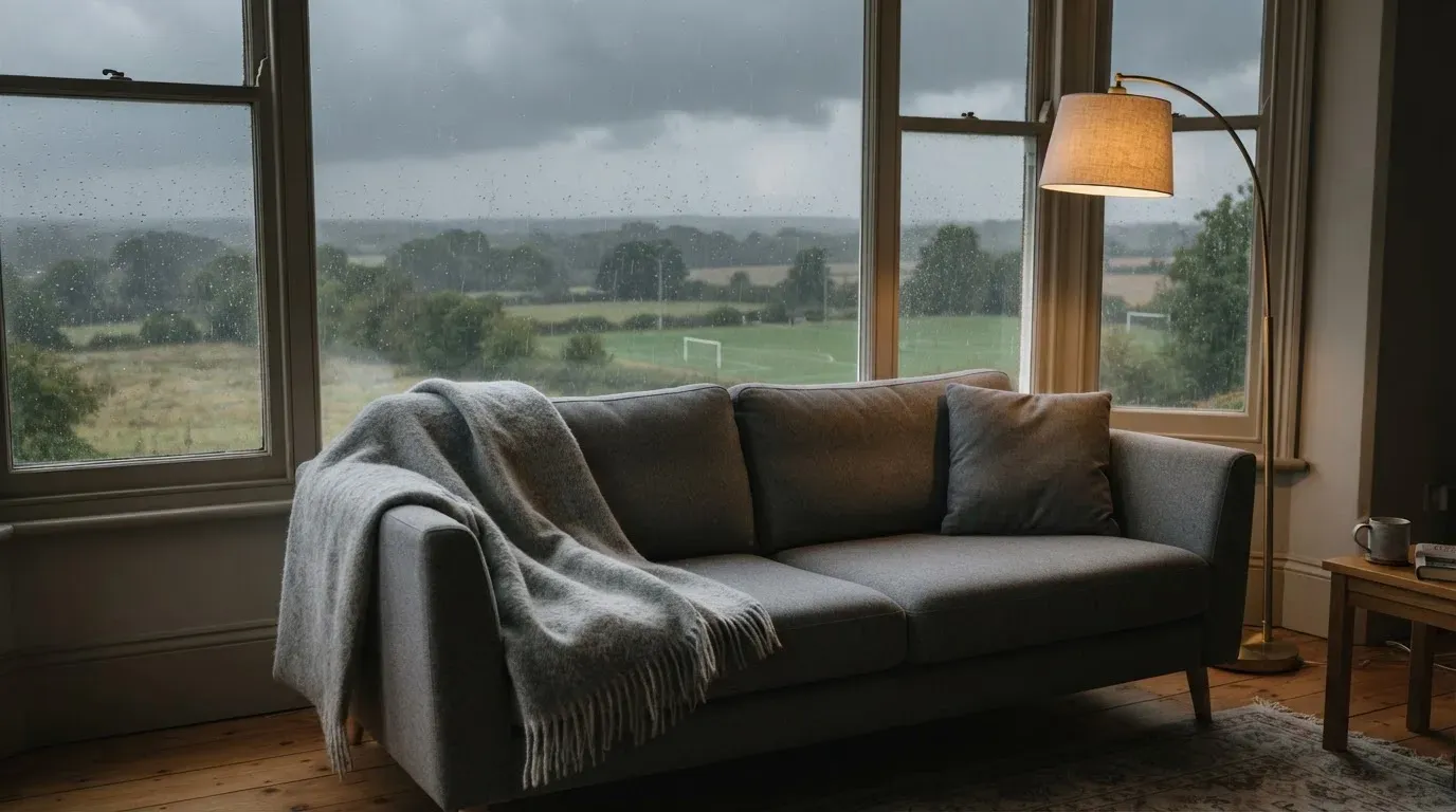 Ein Sofa mit Wolldecke steht vor einem grossen Fenster, draussen Regen und in der Ferne ein Fussballplatz.