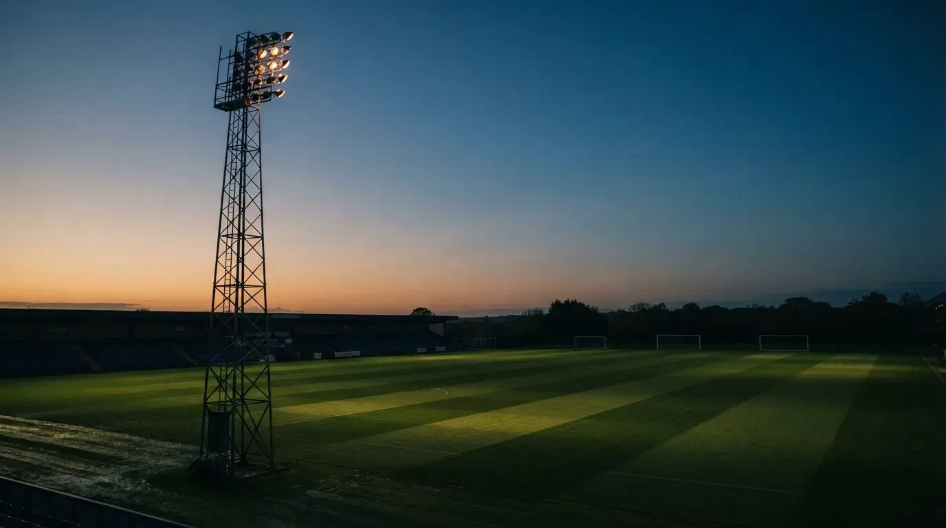Ein Flutlichtmast ragt in den Abendhimmel, die Rasenfläche darunter liegt leer und dunkelgrün da.