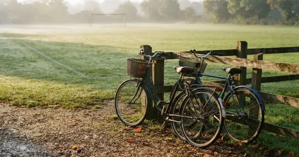 Drei Fahrräder lehnen nebeneinander an einem Zaun vor einem Sportplatz, dahinter ein grüner Rasen im Dunst.