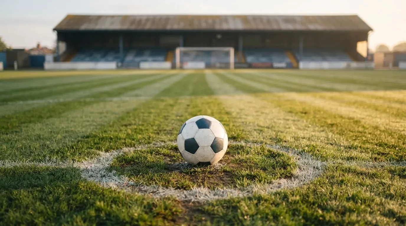 Ein Fussball liegt auf dem Anstosspunkt eines leeren Rasenplatzes im frühen Morgenlicht, dahinter erhebt sich eine menschenleere Tribüne.