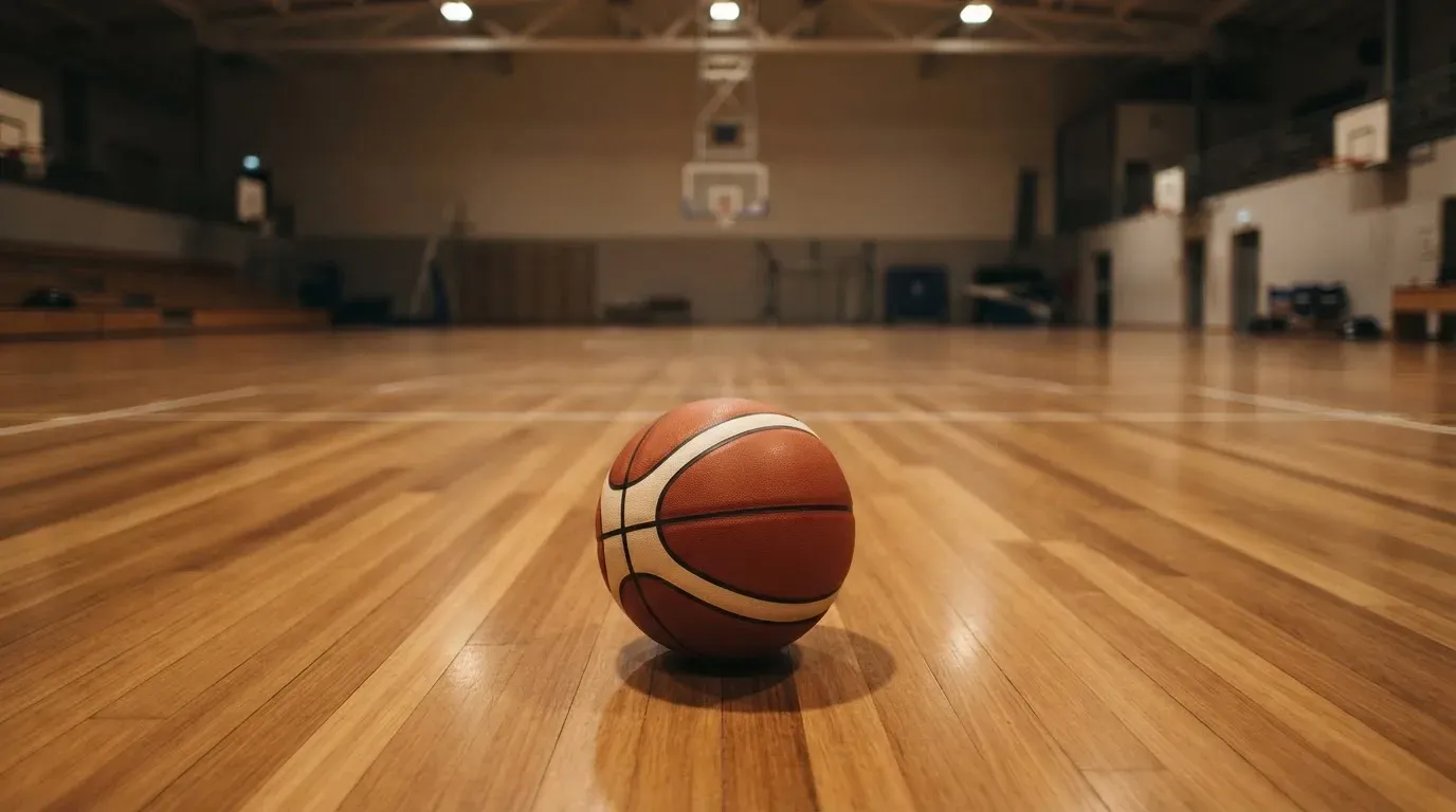Ein Basketball liegt auf dem Parkett einer leeren Halle, von den Deckenlampen fällt gleichmässiges Licht auf den Boden.