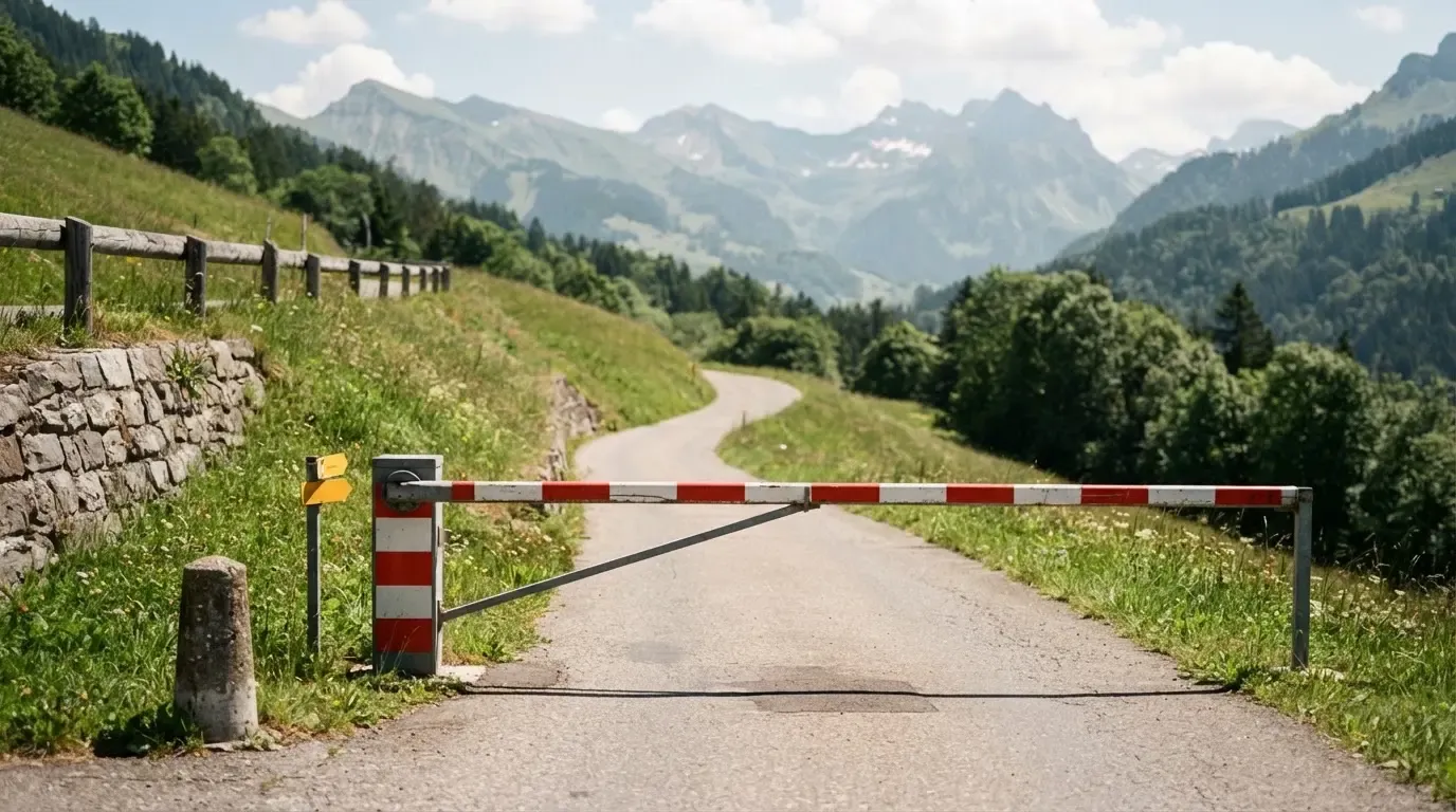 Ein Schlagbaum sperrt eine Landstrasse in den Bergen, dahinter windet sich der Weg zwischen grünen Hängen.