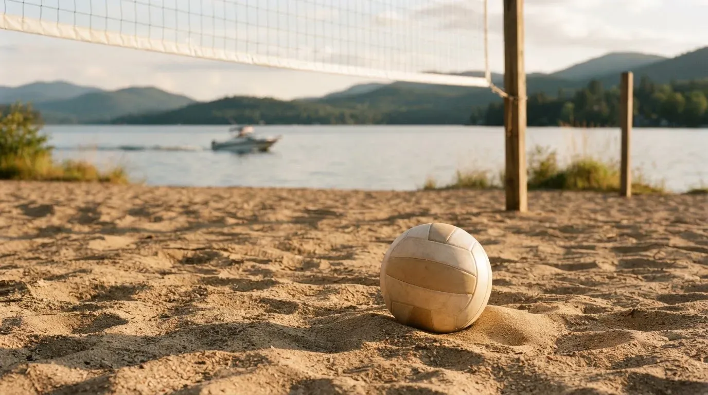Ein Volleyball liegt im Sand eines Spielfelds am Seeufer, im Hintergrund gleitet ein Boot über das Wasser.