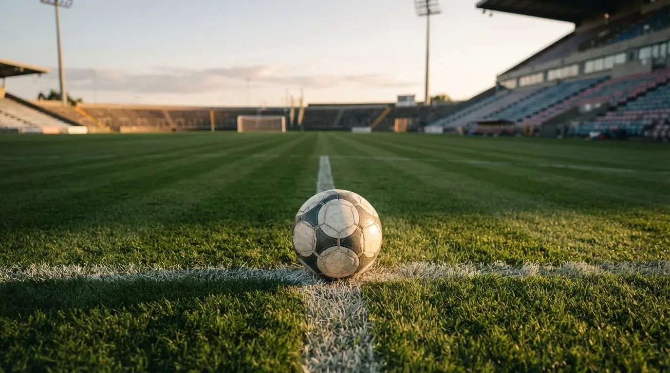 Ein Fussball liegt auf dem Mittelkreis eines leeren Rasenplatzes, dahinter ragen leere Tribünenreihen in den Abendhimmel.