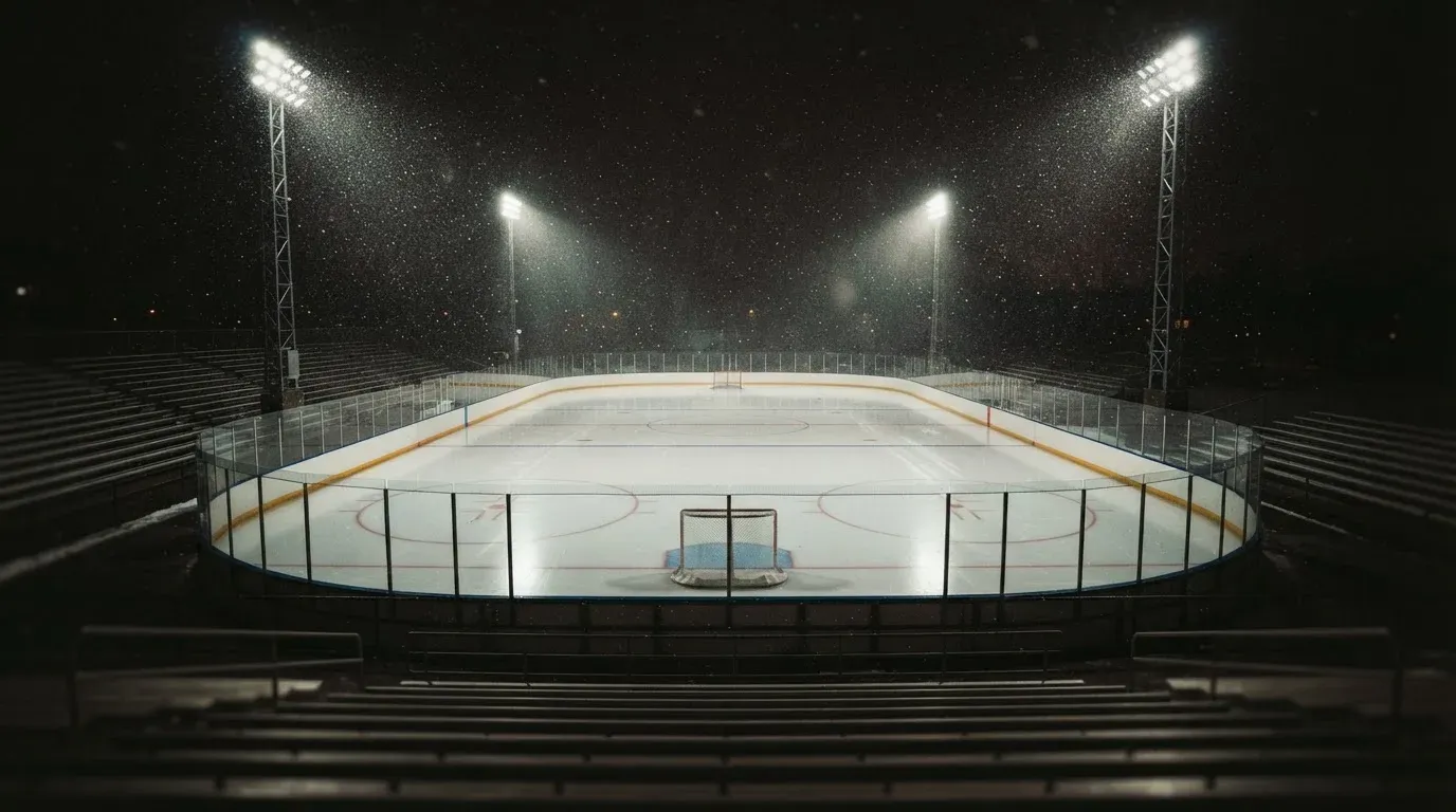 Eine Eishockeybahn liegt unter Flutlicht aus der Ferne betrachtet, leichter Schneefall treibt durch den Lichtkegel über dem Eis.