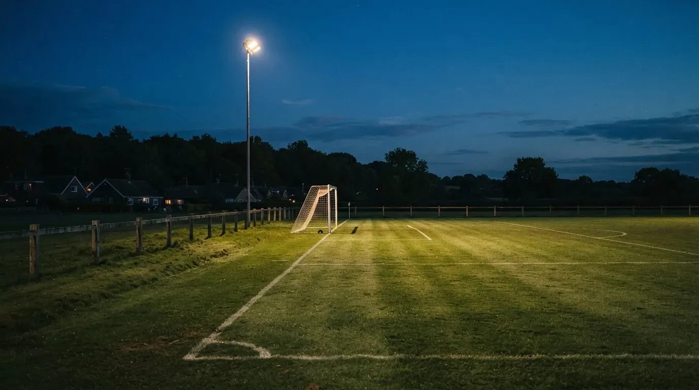 Ein kleiner Dorf-Fussballplatz liegt in der Dämmerung unter einem hohen Flutlichtmast, am Spielfeldrand steht ein Holzzaun.