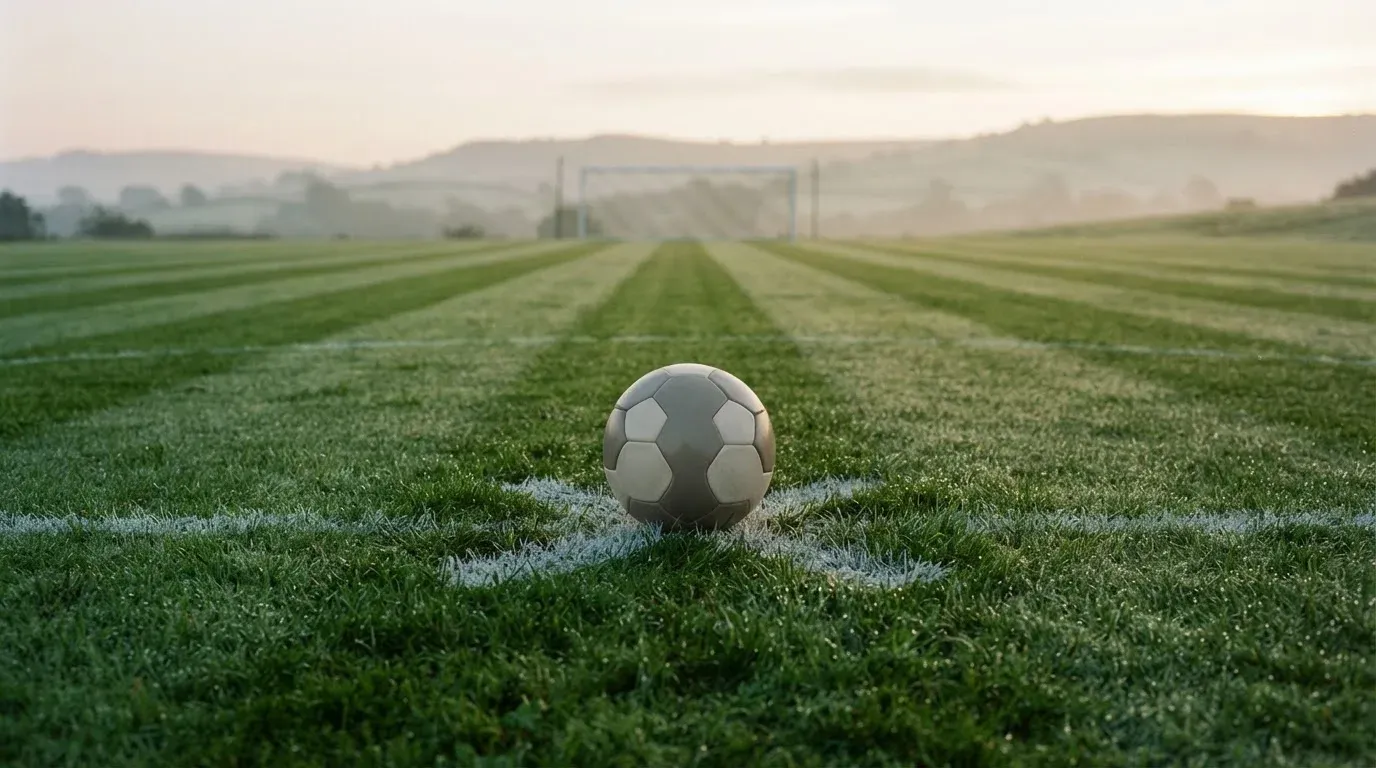 Ein Fussball liegt am frühen Morgen auf dem Anstosspunkt eines taunassen Rasens, im Hintergrund erheben sich Hügel im Dunst.