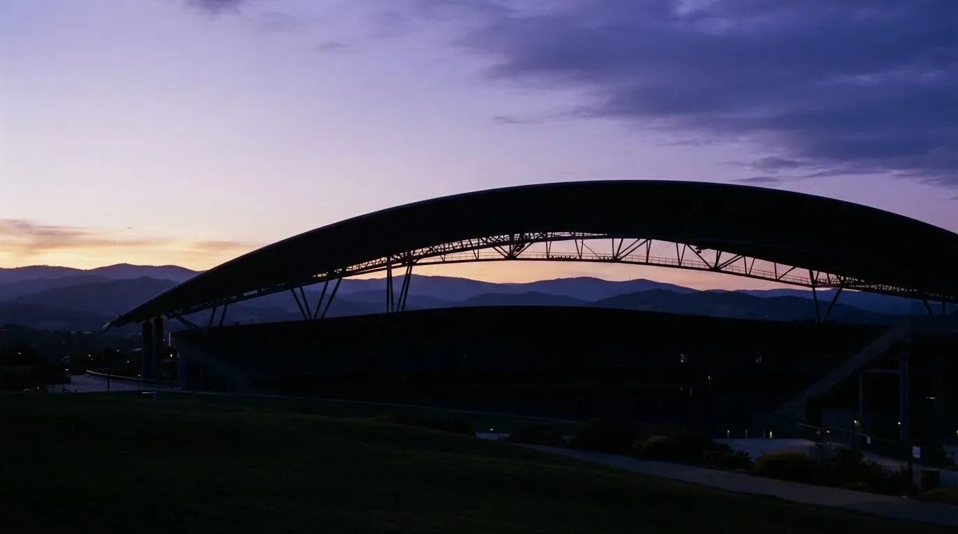 Die Silhouette eines Stadiondachs vor dem violetten Abendhimmel, dahinter eine Hügelkette.