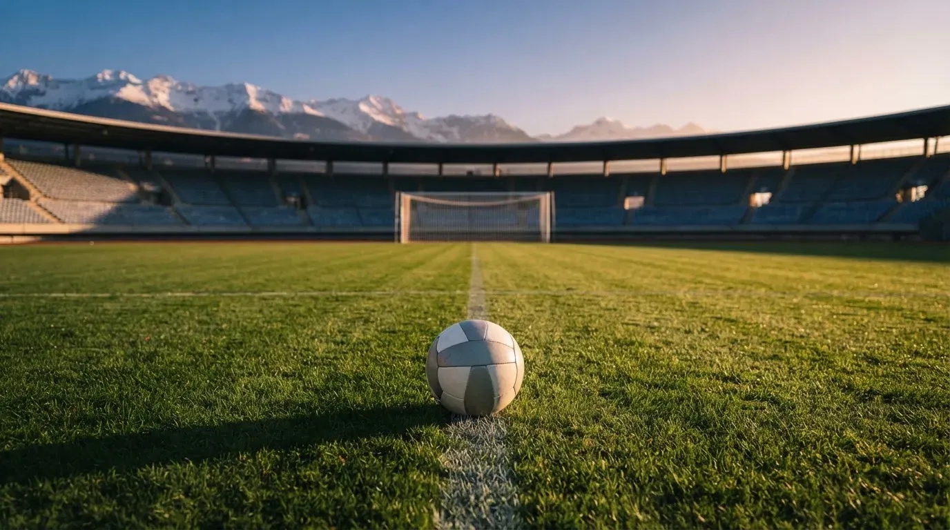 Ein Fussball liegt auf dem Rasen eines leeren Stadions am frühen Abend, dahinter erheben sich schneebedeckte Berge unter klarem Himmel.