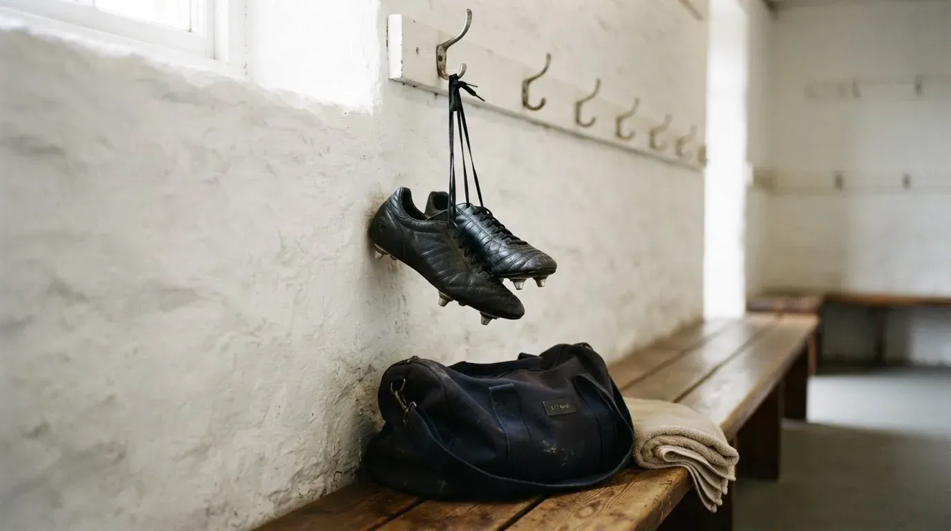 Ein Paar Fussballschuhe hängt an einem Haken in einer ruhigen Umkleide, darunter steht eine Sporttasche auf einer Holzbank.