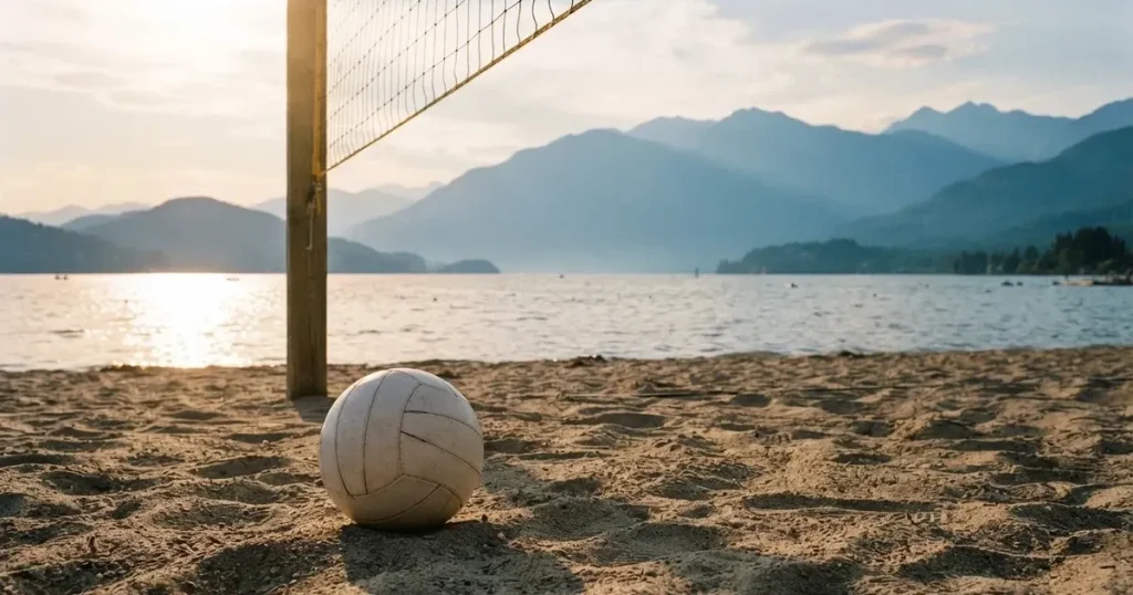 Ein Volleyball liegt am Fuss des Netzes auf Sand, im Hintergrund glitzert ein See und ein Bergzug steht im Dunst.