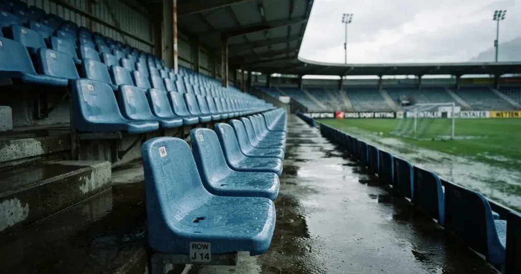 Eine Reihe leerer Sitzschalen auf der Tribüne eines kleinen Stadions, auf den Stufen glänzt Regenwasser.
