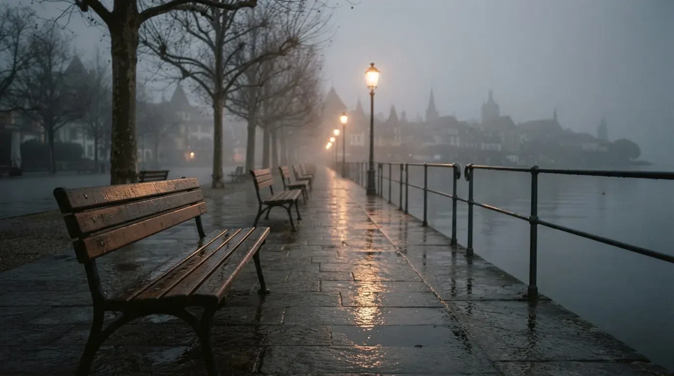 Eine Seepromenade mit leeren Bänken an einem nebligen Morgen, im Wasser spiegeln sich die Lichter der Uferlaternen.
