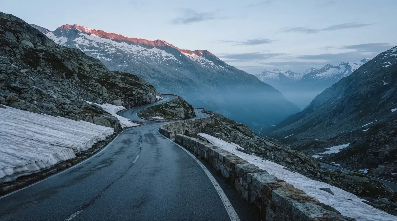 Ein Bergpass in den Schweizer Alpen mit leerer Strasse und Schneeresten am Rand im ersten Frühlicht.