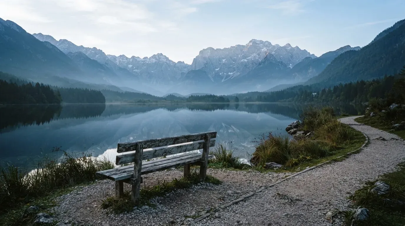 Eine Holzbank an einem Seeufer mit Blick auf eine Bergkette im Morgendunst, das Wasser ist spiegelglatt.