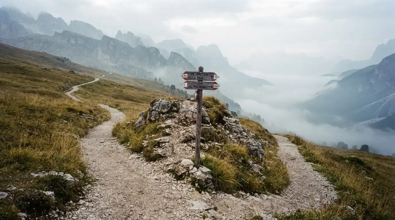 Eine Weggabelung auf einer Alpweide, zwei Pfade teilen sich zwischen Steinen, im Tal steigt Nebel auf.