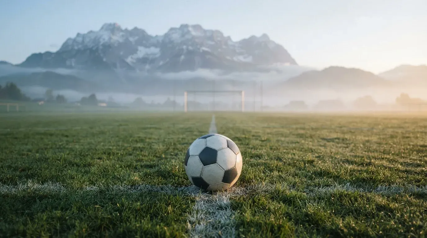 Ein einzelner Fussball auf dem Mittelpunkt eines leeren Rasens im Morgennebel, hinter dem Tor ragen Berge auf