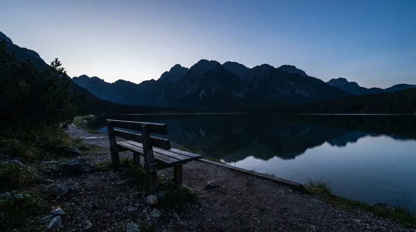 Eine leere Holzbank am Seeufer in der Dämmerung, im Hintergrund dunkle Berge