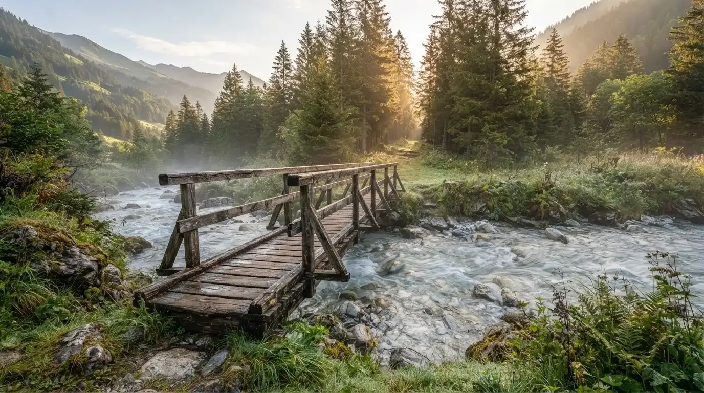 Eine alte Holzbrücke führt über einen Bergbach im Morgenlicht, Tannen säumen das Ufer