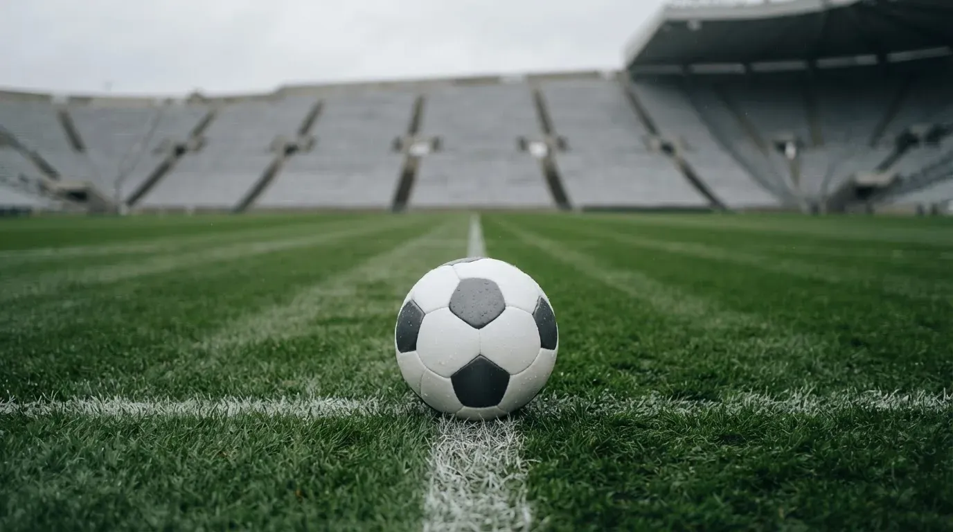 Ein Fussball liegt auf dem Anstosspunkt eines nassen Rasens, dahinter eine leere Tribüne unter grauem Himmel.