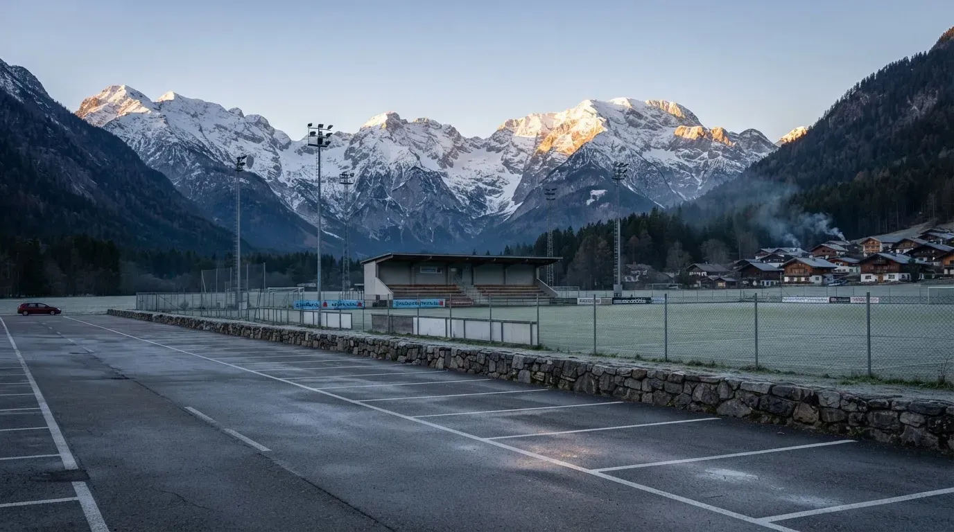Schneebedeckte Berge ragen hinter einem kleinen Stadion in einem Alpental auf, im Vordergrund ein leerer Parkplatz am Morgen.