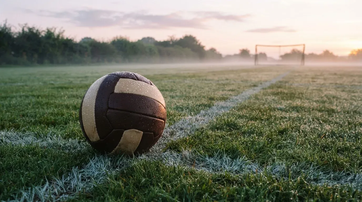 Ein Fussball liegt im Tau auf der Mittellinie eines Dorfplatzes bei Sonnenaufgang, der Rasen glitzert.