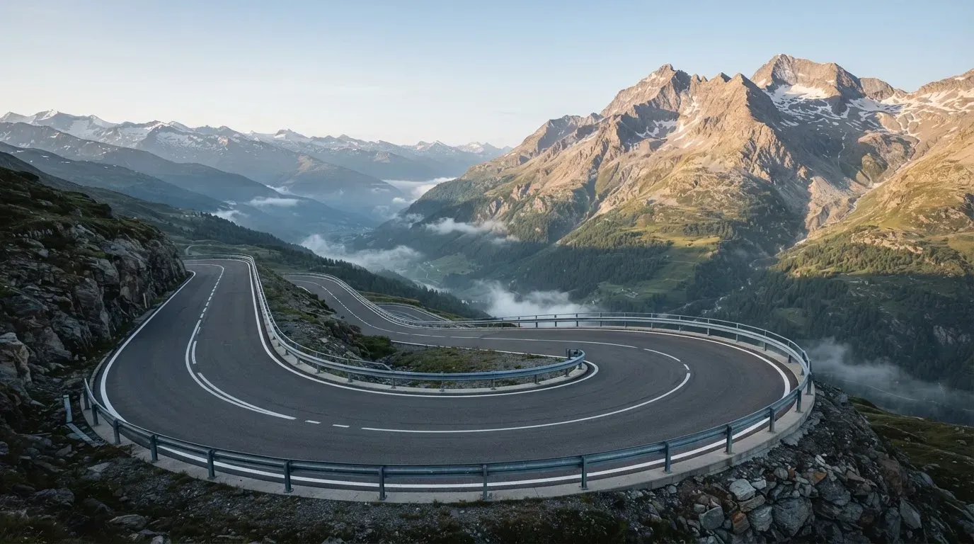 Eine Passstrasse in den Alpen mit enger Kurve und Leitplanke, darüber ein klarer Morgenhimmel und einzelne Nebelschwaden im Tal.