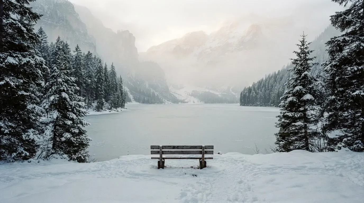 Ein zugefrorener Bergsee im Morgendunst, am Ufer steht eine einsame Holzbank zwischen verschneiten Tannen.