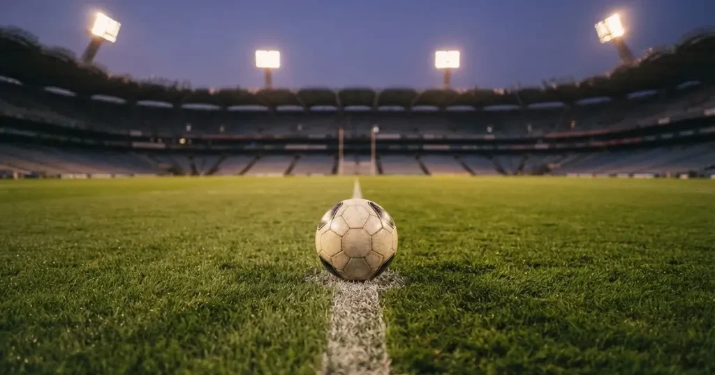 Ein Fussball liegt auf dem Anstosspunkt eines leeren Rasenplatzes, dahinter ragen die Ränge eines grossen Stadions in der Abenddämmerung auf.