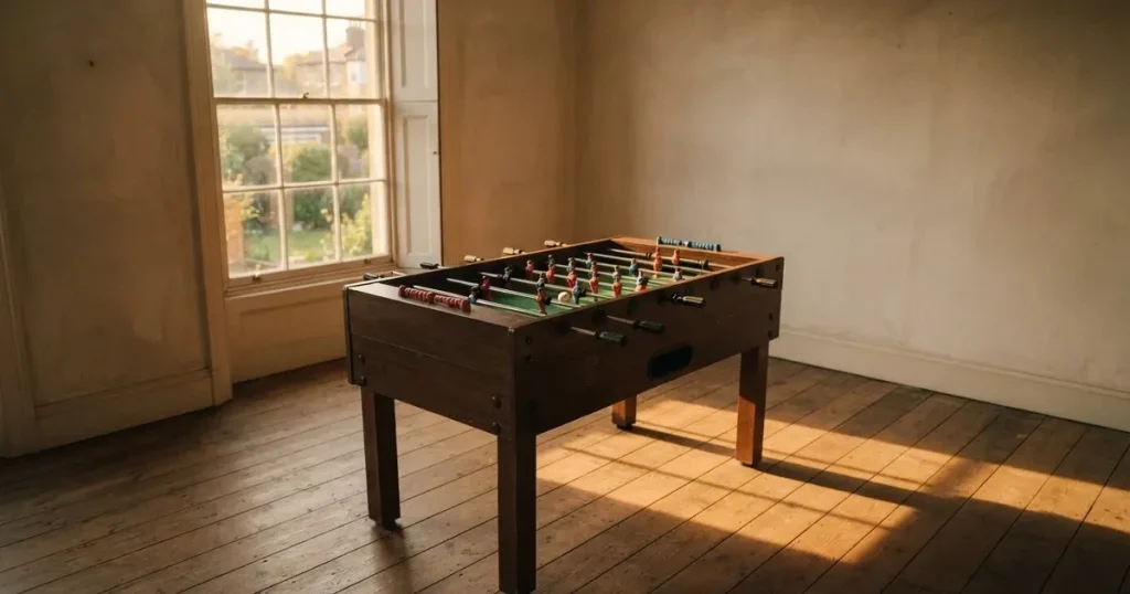 Ein Tischfussballtisch steht in einem leeren Raum, auf dem grünen Spielfeld liegt eine einzelne Kugel im Licht eines Fensters.