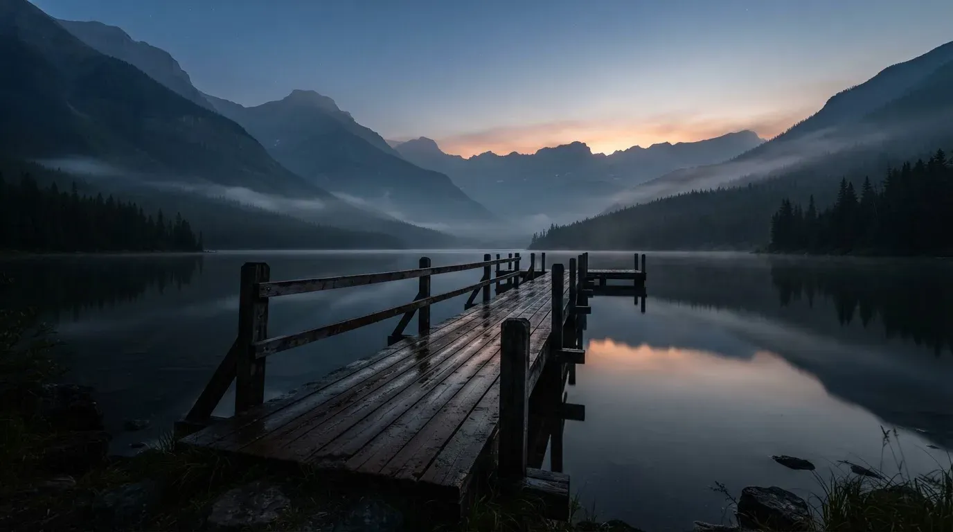 Ein Bergsee in der Dämmerung mit einem leeren Holzsteg, die Wasseroberfläche ist ruhig und Nebel zieht über die Hänge.