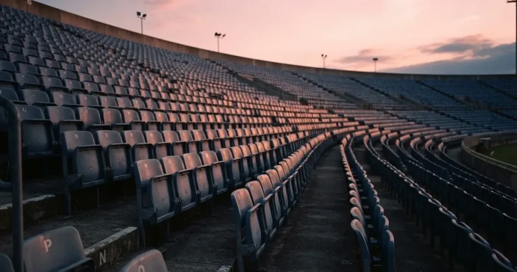 Eine Reihe leerer Sitzplätze in einem Stadion, die Sessel in gleichmässigem Abstand, über den Rängen ein Abendhimmel in Rosa.