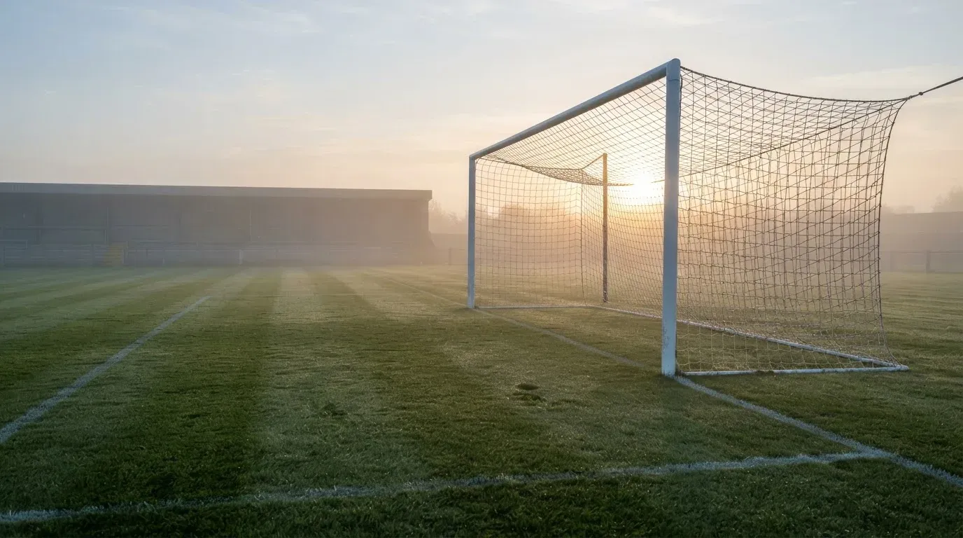 Ein Torpfosten mit Netz steht im Gegenlicht auf einem leeren Fussballplatz, über dem Rasen liegt Nebel.