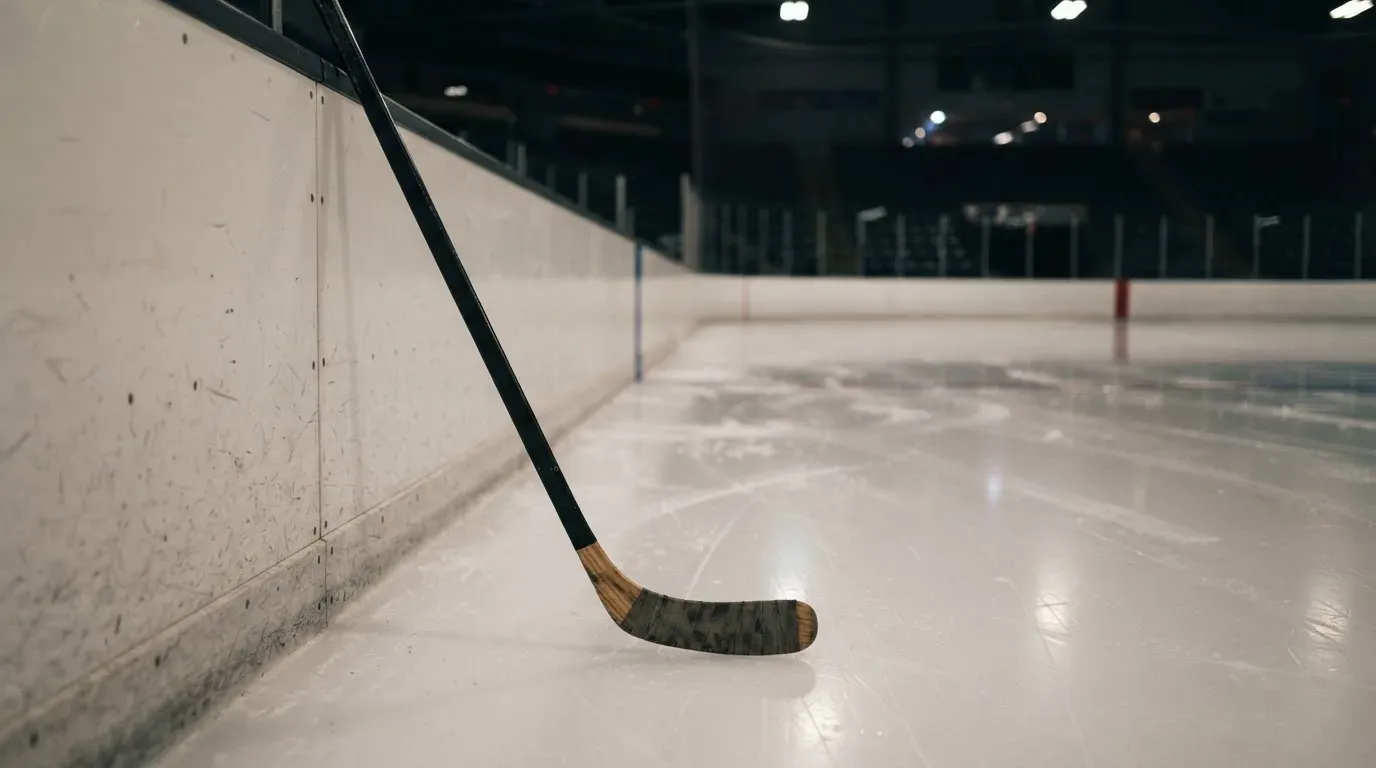 Ein Eishockeyschläger lehnt an der Bande einer leeren Halle, auf dem Eis spiegeln sich die Deckenlampen.