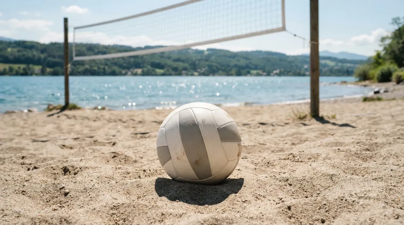 Ein Volleyball liegt auf dem Sand eines Strandfeldes am Seeufer, im Hintergrund glitzert das Wasser.