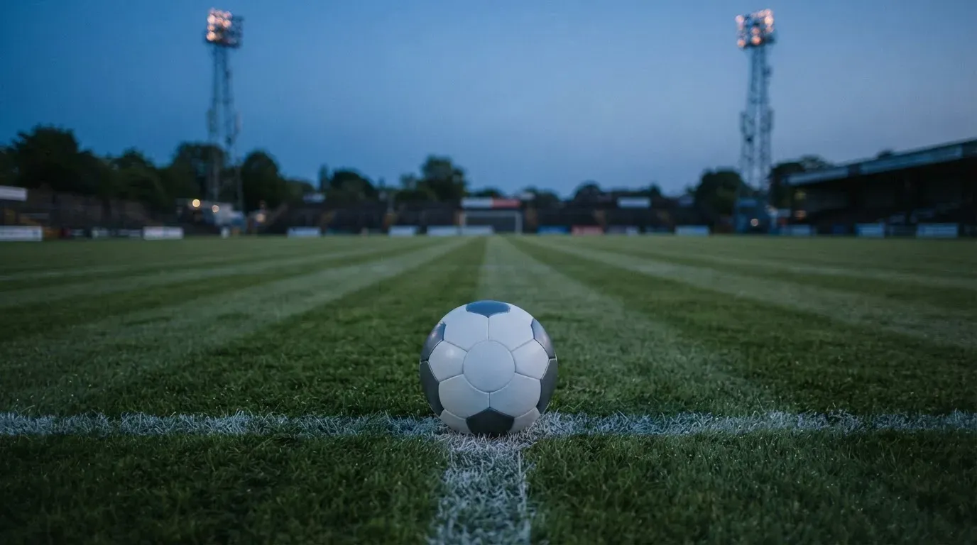 Ein Fussball liegt auf dem Mittelpunkt eines leeren Rasenplatzes, im Hintergrund ragen Flutlichtmasten in die Abenddämmerung