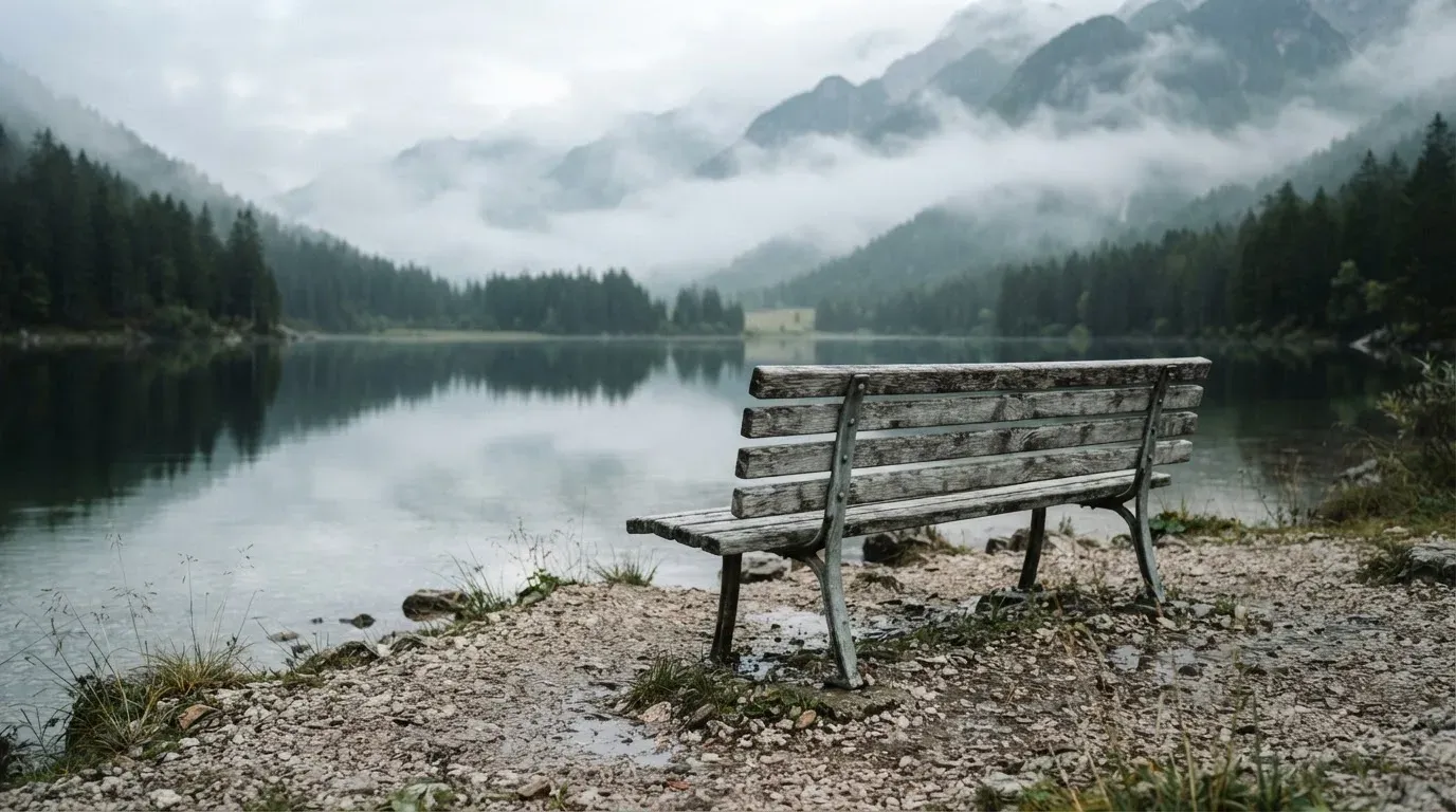 Eine Holzbank am Ufer eines ruhigen Bergsees, am gegenüberliegenden Ufer steigen dunkle Hänge in den Frühnebel