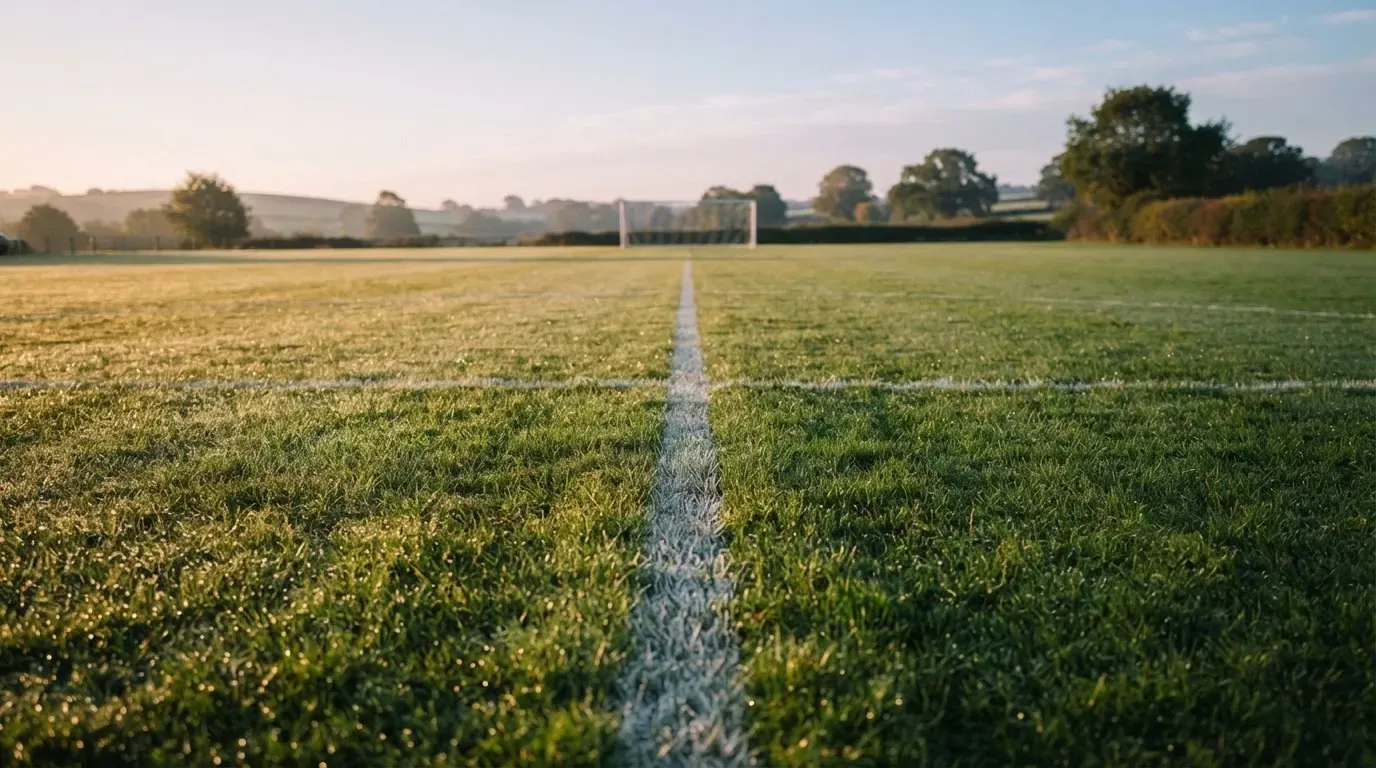 Ein Rasenspielfeld bei Sonnenaufgang, die Mittellinie verläuft quer durchs Bild und Tau glitzert auf dem Gras.