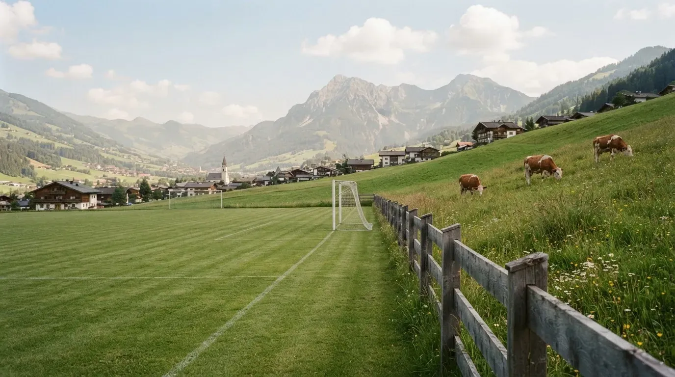 Ein Dorffussballplatz mit Holzzaun am Hang, dahinter grasen Kühe auf einer Weide unter hohen Wolken.