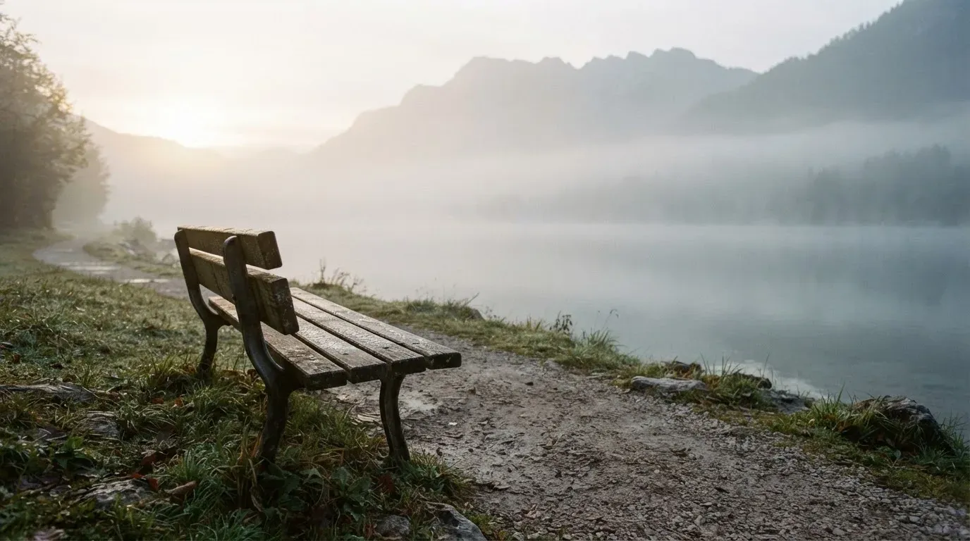 Eine leere Parkbank am Seeufer im Morgennebel, das Wasser liegt still und die Bergkette ist nur zu erahnen.