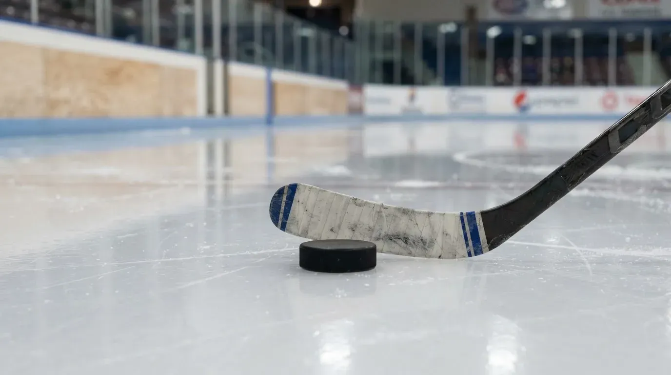 Ein Eishockeypuck und ein Schläger liegen auf einer glänzenden Eisfläche, im Hintergrund verschwimmen leere Banden.