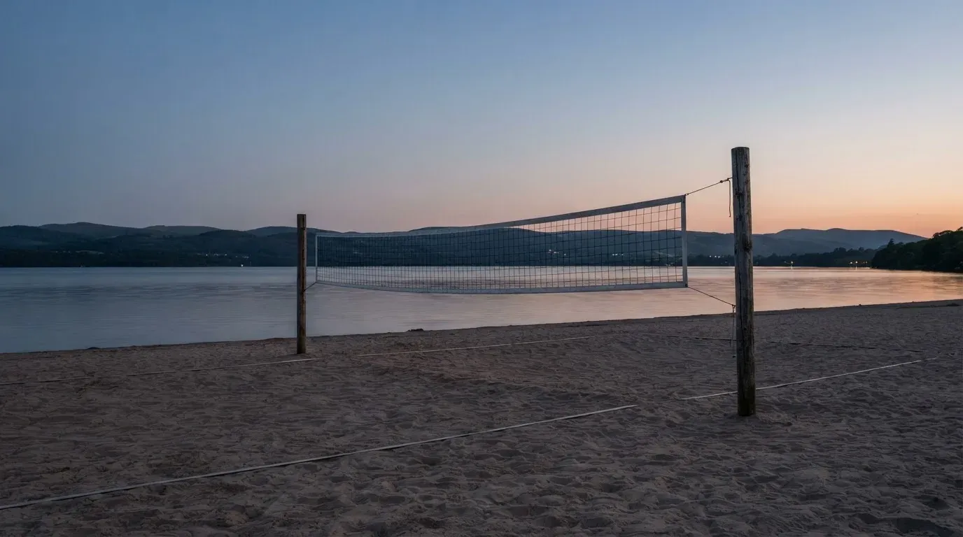 Ein Volleyballnetz steht an einem Seeufer in der Abenddämmerung, der Sand ist leer und das Wasser glatt.