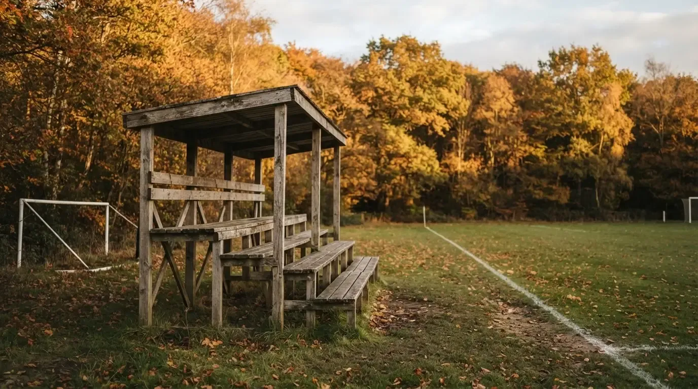 Eine leere Holztribüne steht am Waldrand neben einem kleinen Sportplatz im Herbstlicht.