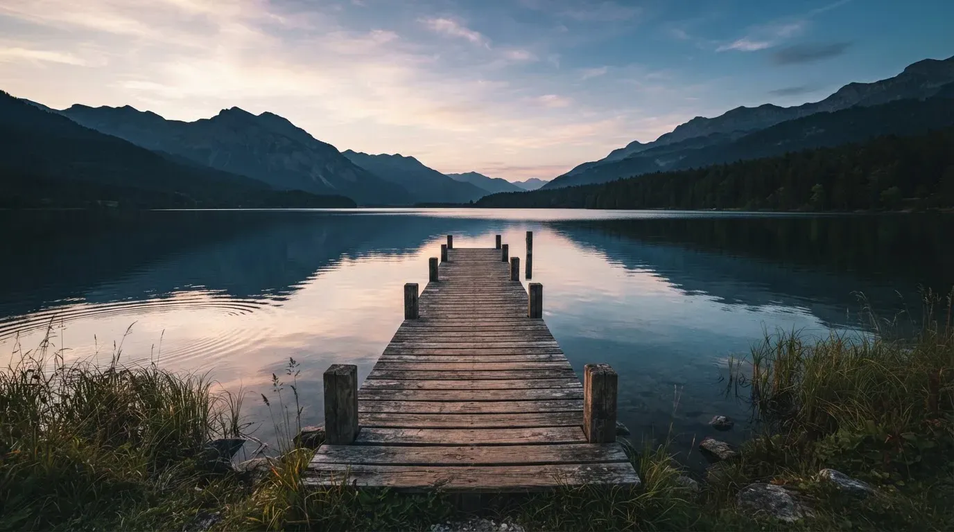 Ein leerer Holzsteg ragt am Abend in einen ruhigen Bergsee, das Wasser spiegelt den Himmel.