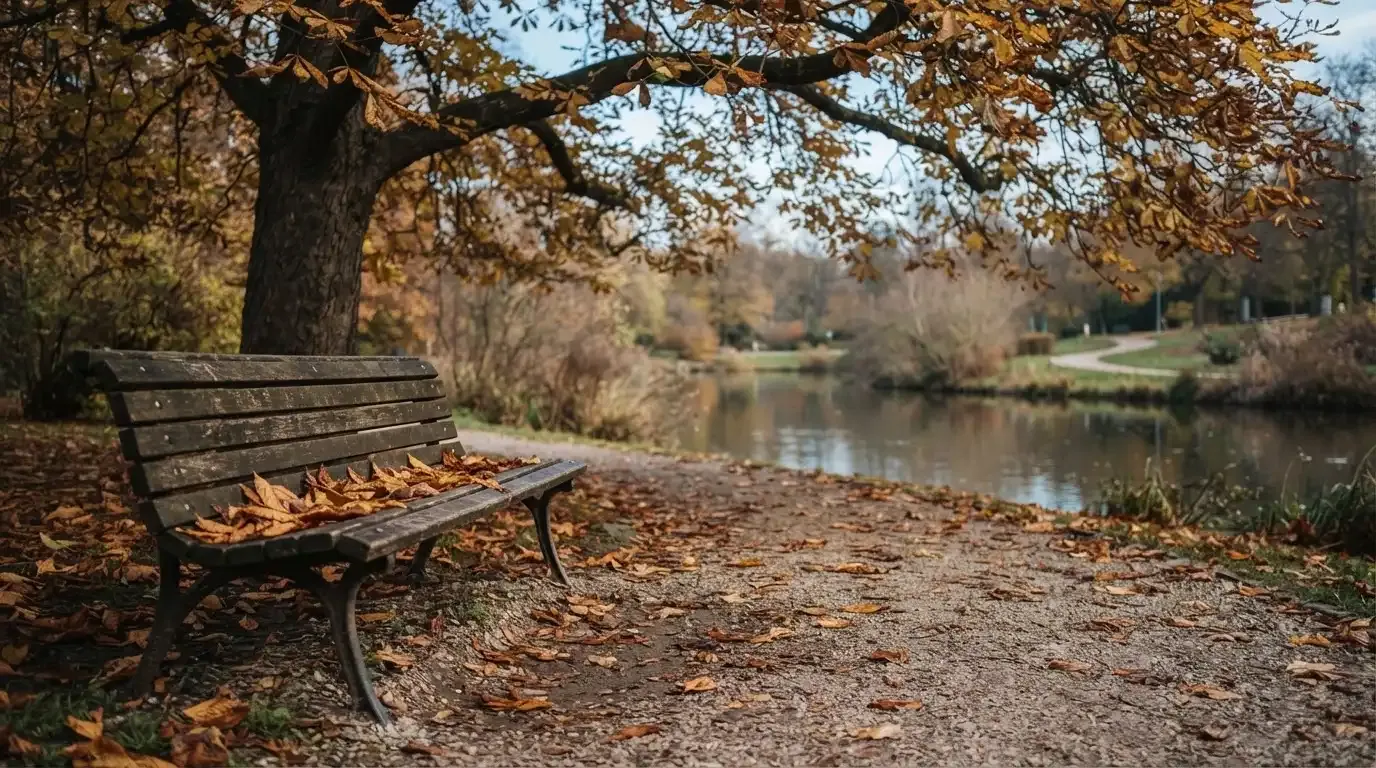 Eine Parkbank unter einer Kastanie, Herbstlaub bedeckt den Kiesweg, im Hintergrund ein stiller Teich.