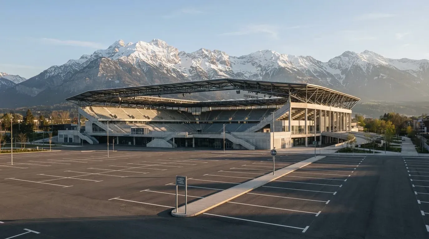 Hinter einem leeren Stadion ragen verschneite Berggipfel in den klaren Morgenhimmel, davor ein Parkplatz im ersten Sonnenlicht.