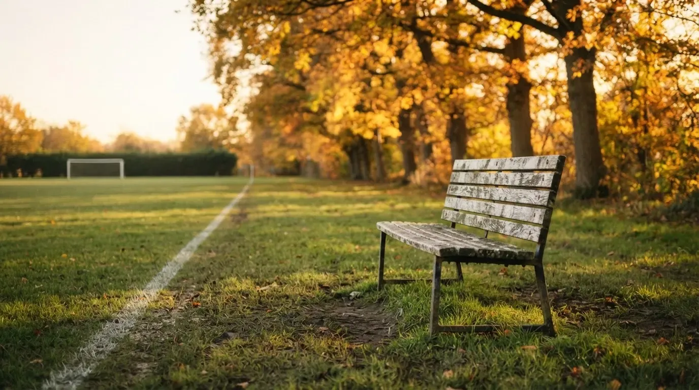 Eine hölzerne Bank steht an der Seitenlinie eines leeren Sportplatzes, dahinter Bäume im Herbstlicht.